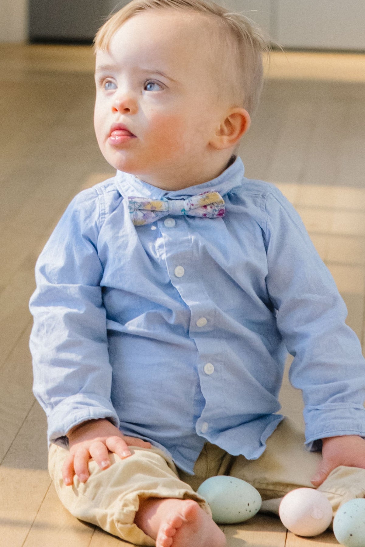 A young child sits on a wooden floor next to pastel-colored eggs, wearing Ivy City Co’s “Ties in Pretty Plumeria” paired with a light blue shirt and looking up with a thoughtful expression—perfectly dressed for any spring celebration.