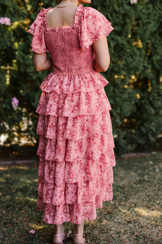 A woman stands outdoors on grass, wearing the Ivy City Co Grace Dress in Desert Heritage Rose. The image showcases the ruffled details, flutter sleeves, and layered fabric of this maternity-friendly style from behind, with lush green foliage in the background.