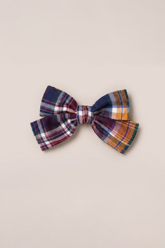 The Hair Bows in Midnight Plaid by Ivy City Co—a large accessory adorned with a classic plaid pattern in navy blue, burgundy, white, yellow, and orange—rests elegantly on a plain beige background, capturing timeless Ivy City charm.
