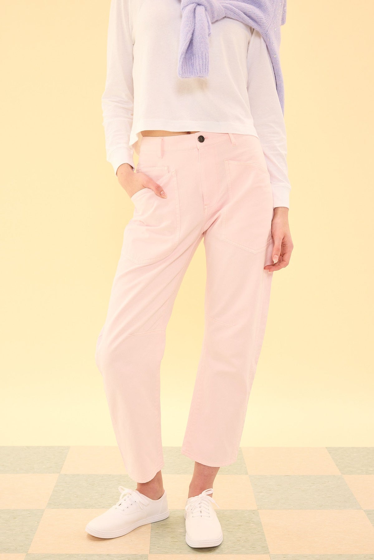 A person wearing the Amanda Pants in Light Pink by Ivy City Co, a white long-sleeve shirt, a lavender sweater draped over their shoulders, and white sneakers stands on a checkered floor against a pale yellow background. The Amanda Pants in Light Pink bring a fresh pop of color to this effortlessly chic ensemble.