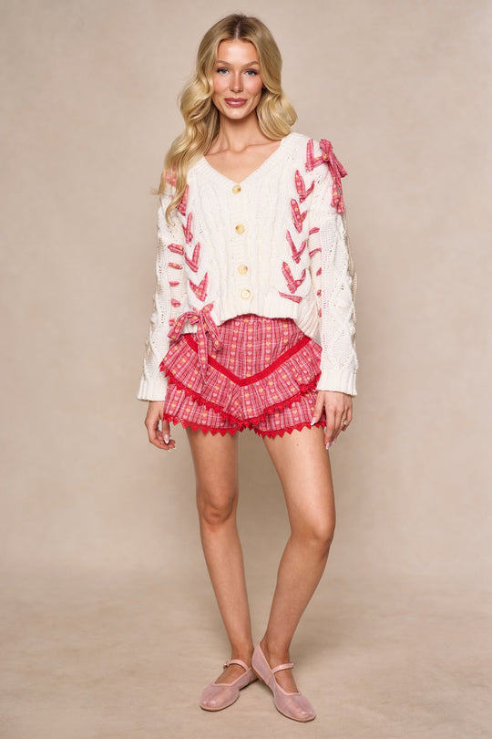 Woman with long blonde hair wears the Ivy City Co Agatha Sweater in Ivory, featuring pink ribbon details, paired with pink plaid ruffle shorts and pink Mary Jane shoes. She stands and smiles against a neutral background.