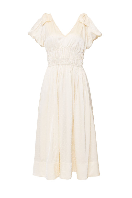 The Zella Bow Dress in Ivory by Ivy City Co features a cream-colored, short-sleeve midi silhouette with a V-neckline, gathered waist, and subtle textured dot pattern. This elegant dress is an ideal choice for a rehearsal dinner or can be styled as a modern bridal look.