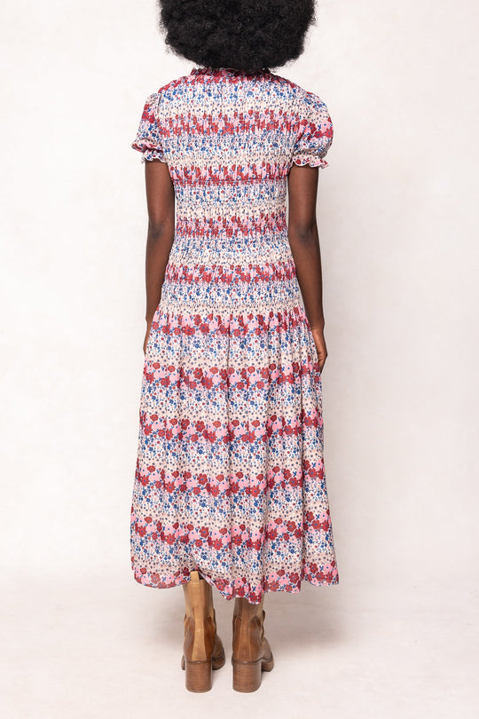 A woman with curly hair stands facing away, wearing Ivy City Co’s Hayden Dress in Lush Garden—a short-sleeve, ankle-length floral midi dress featuring a smocked bodice—paired with brown ankle boots. This look is both stylish and maternity friendly.
