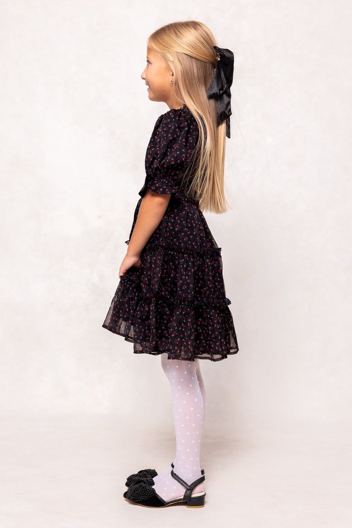 A young girl, smiling in profile against a light background, wears the Ivy City Co Mini Madeline Dress in Black Ditsy Floral—styled with white polka dot tights, black bow-adorned shoes, and a large black hair ribbon.