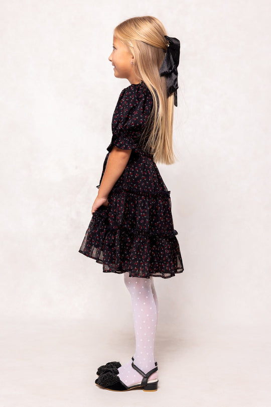 A young girl, smiling in profile against a light background, wears the Ivy City Co Mini Madeline Dress in Black Ditsy Floral—styled with white polka dot tights, black bow-adorned shoes, and a large black hair ribbon.