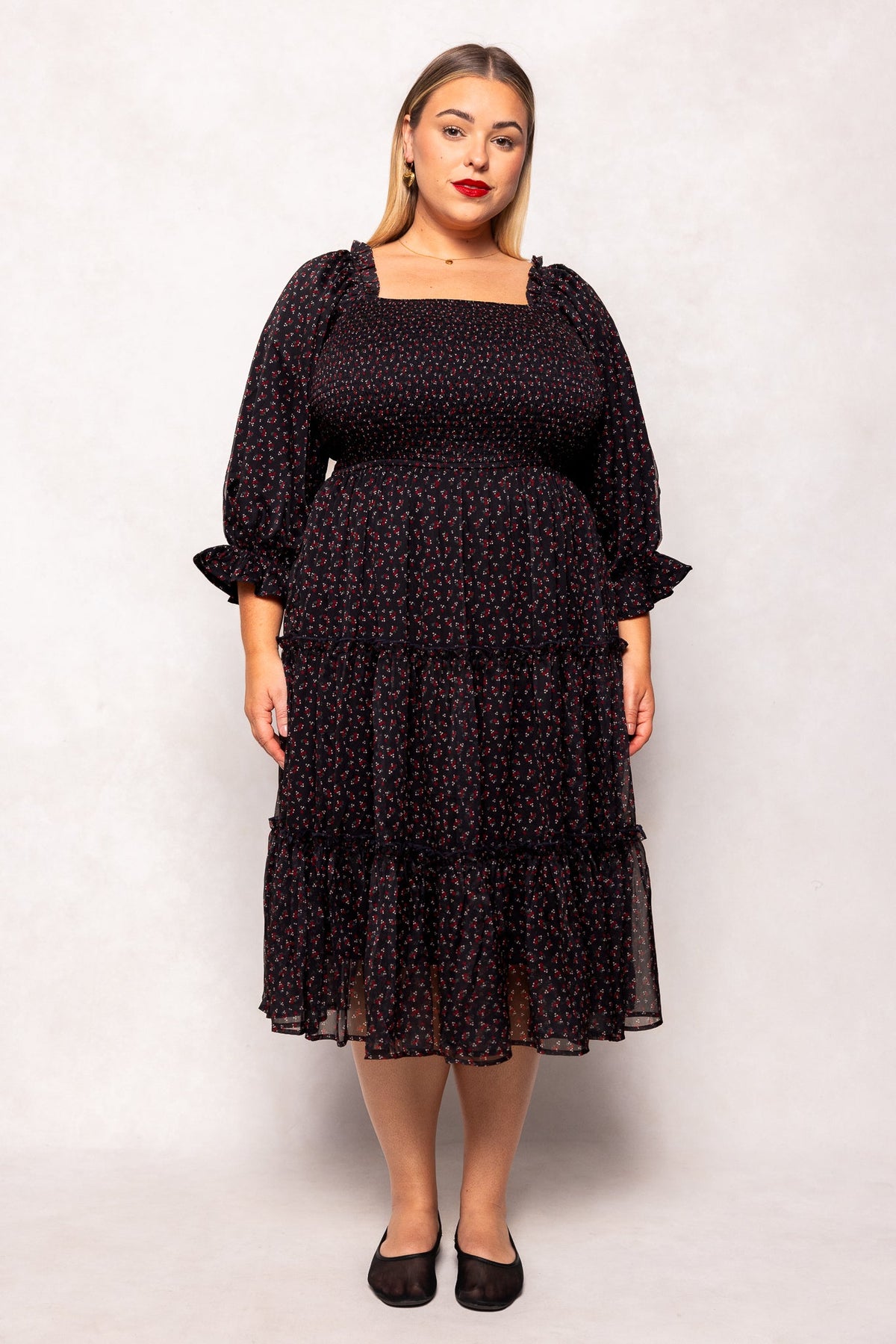 A woman stands facing forward against a light backdrop, wearing the Ivy City Co Madeline Dress in Black Ditsy Floral. The maternity-friendly design features long sleeves, a square neckline, and a ruffled hem that drapes gracefully over her figure. She has straight, shoulder-length hair and pairs the dress with black flats.