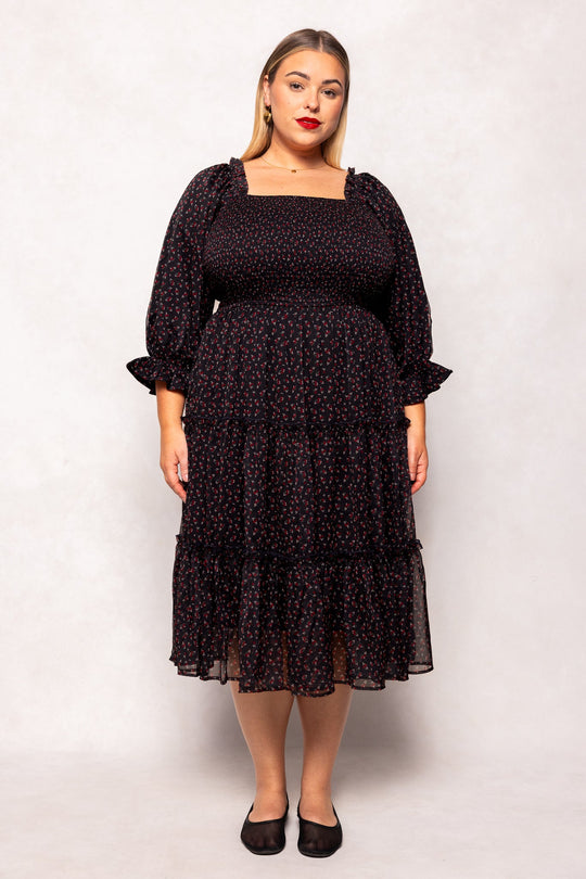 A woman stands facing forward against a light backdrop, wearing the Ivy City Co Madeline Dress in Black Ditsy Floral. The maternity-friendly design features long sleeves, a square neckline, and a ruffled hem that drapes gracefully over her figure. She has straight, shoulder-length hair and pairs the dress with black flats.
