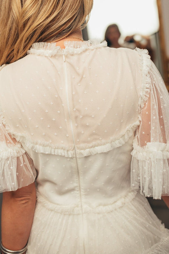 A woman is seen from the back wearing the Ivy City Co Whimsical Dress in White, featuring sheer tulle with ruffled sleeves and delicate polka dot details. Her loose hair enhances the dreamy charm of her look, while another person appears blurred in the background.