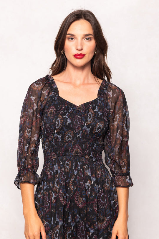 A woman with long brown hair wears the Maddison Dress in Enchanted Floral by Ivy City Co—a sheer, dark floral design featuring three-quarter sleeves. She stands facing forward against a plain light background, her neutral expression and bold red lipstick drawing attention to the dress’s effortless elegance. This look is maternity friendly, offering both comfort and sophistication.