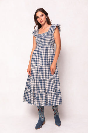A woman wears the Ivy City Co Hailey Dress in Indigo Gingham—a midi nursing-friendly dress with a smocked bodice and tiered skirt—paired with floral embroidered blue boots, standing against a light background.