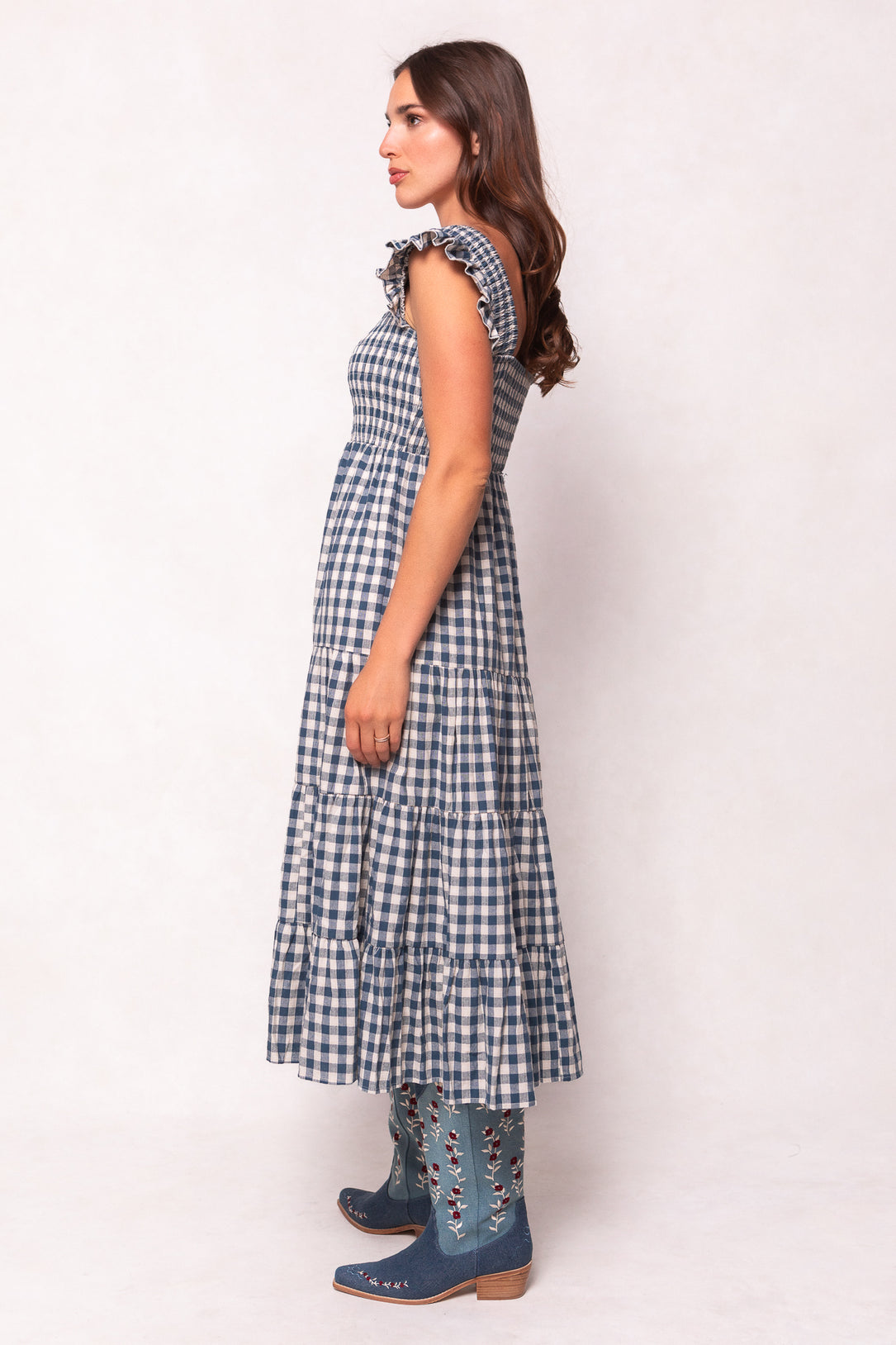 A woman with long, wavy brown hair stands in profile wearing Ivy City Cos Hailey Dress in Indigo Gingham—a sleeveless, maternity-friendly style with ruffled shoulders and a tiered skirt—paired with blue embroidered knee-high boots.
