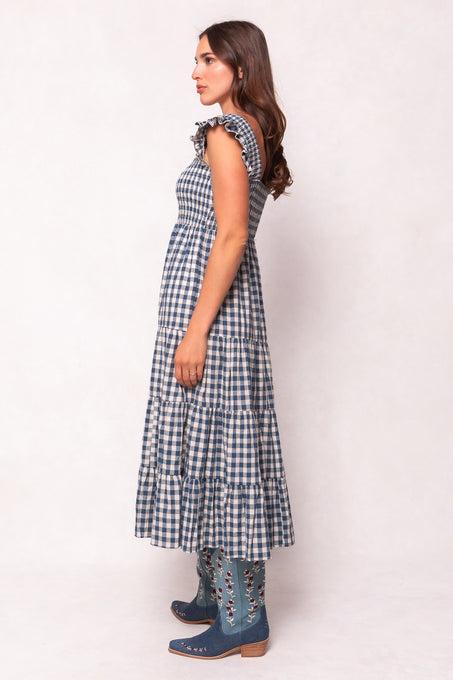 A woman with long, wavy brown hair stands in profile wearing Ivy City Cos Hailey Dress in Indigo Gingham—a sleeveless, maternity-friendly style with ruffled shoulders and a tiered skirt—paired with blue embroidered knee-high boots.