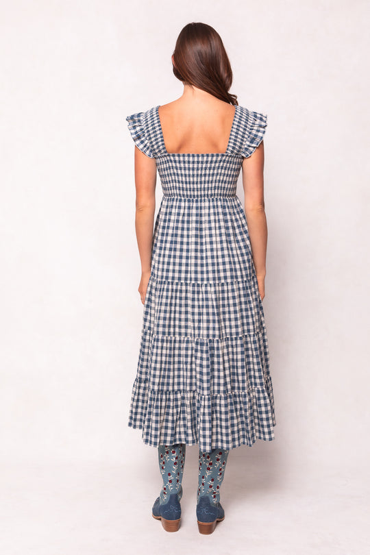 A woman faces away in the Ivy City Co Hailey Dress in Indigo Gingham—a maternity and nursing-friendly midi with ruffled sleeves. Her brown hair falls to one side, and she pairs it with blue, floral-embroidered boots against a light background.