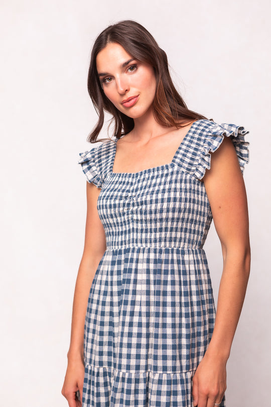 A woman with long brown hair wears the Ivy City Co Hailey Dress in Indigo Gingham, featuring ruffled sleeves, while standing against a plain light background and gazing slightly to the side.