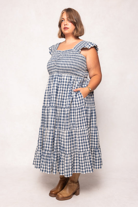A woman in the Ivy City Co Hailey Dress in Indigo Gingham, featuring ruffled sleeves and a nursing-friendly design, stands slightly to the side against a plain light background. She wears brown ankle boots, a watch, and has short hair.