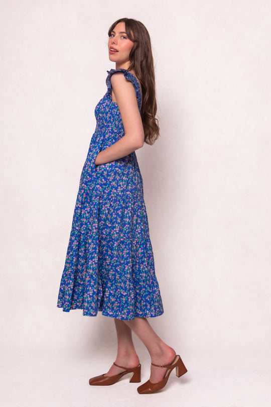 A woman with long brown hair stands sideways in the Ivy City Co Hailey Dress in Allie Blue Floral, featuring ruffled sleeves and pockets. She pairs it with brown heels against a light, plain background.