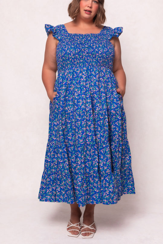 A woman stands against a light background wearing Ivy City Co’s Hailey Dress in Allie Blue Floral, a maternity friendly style with ruffled shoulder straps, paired with white strappy sandals for a fresh, stylish look.