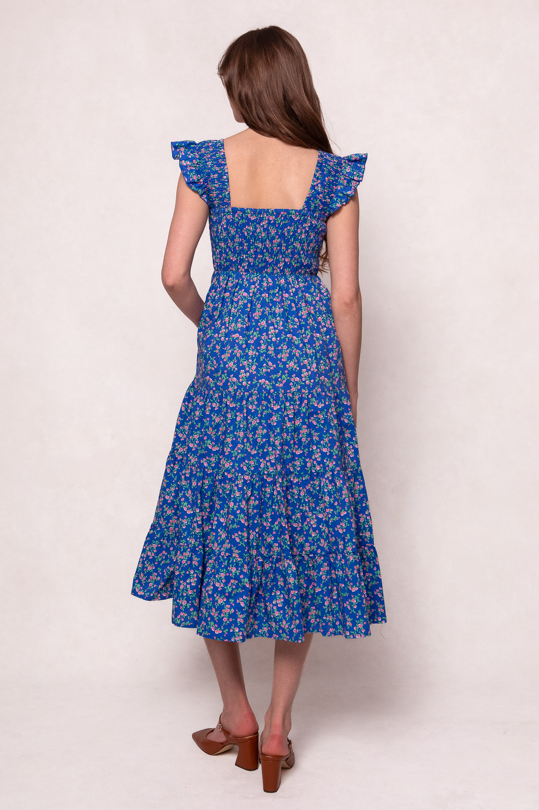 A woman with long brown hair faces away, wearing the Hailey Dress in Allie Blue Floral by Ivy City Co—an ankle-length, maternity-friendly dress with ruffled sleeves—paired with brown heeled sandals against a plain light background.