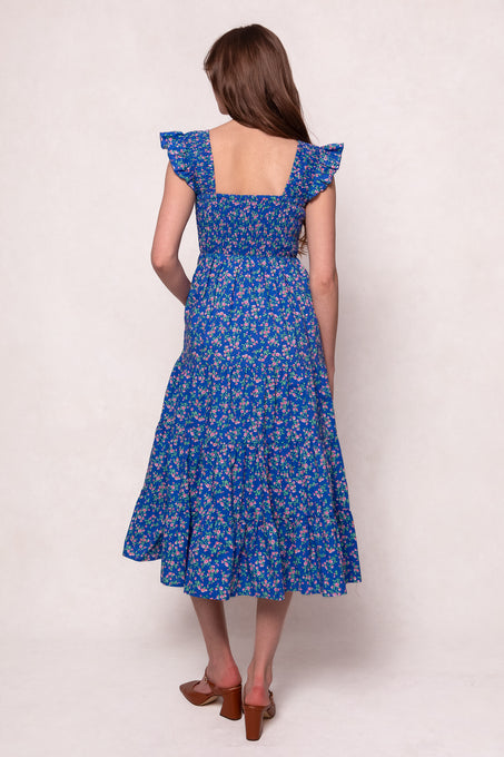 A woman with long brown hair faces away, wearing the Hailey Dress in Allie Blue Floral by Ivy City Co—an ankle-length, maternity-friendly dress with ruffled sleeves—paired with brown heeled sandals against a plain light background.