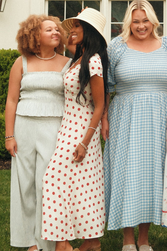 Three women stand outside, smiling. One wears Ivy City Co's Harley Pants in Blue & White Pinstripe, another is in a maternity-friendly white dress with red polka dots and a sunhat, while the third sports a blue gingham dress. They look happy and relaxed.