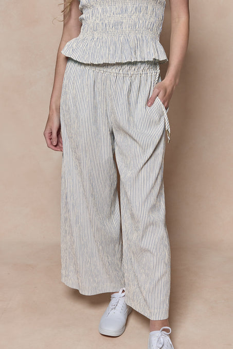 Wearing Ivy City Co's Harley Pants in Blue & White Pinstripe with a matching sleeveless top and classic white sneakers, one hand in their pocket, this beige background look is maternity-friendly.