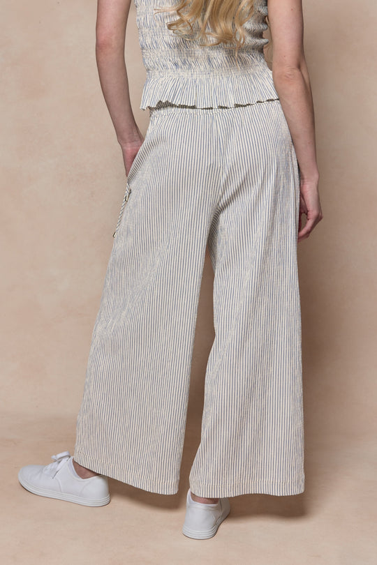 A person with long blonde hair stands facing away, wearing Ivy City Co's Harley Pants in Blue & White Pinstripe, a matching sleeveless top, and white sneakers against a light beige background.