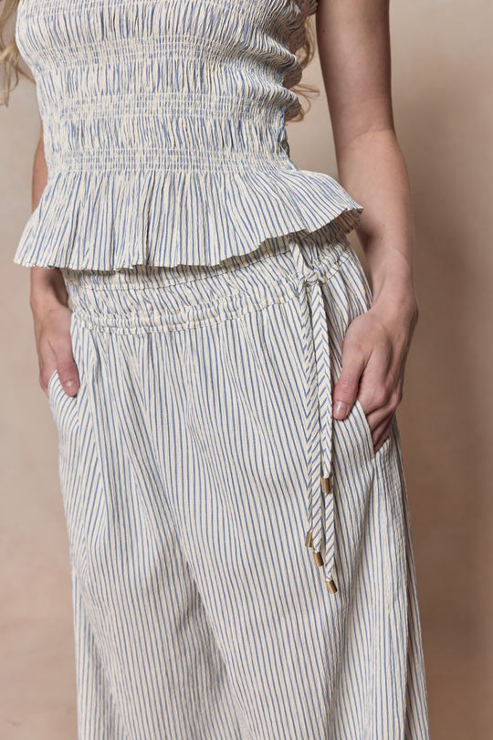 A person wearing Ivy City Co’s Harley Pants in Blue & White Pinstripe with a coordinating, maternity-friendly, ruffled hem sleeveless top stands with hands in pockets against a beige background.