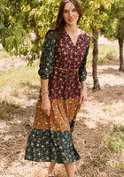 A woman stands outdoors among trees, wearing the Harlowe Dress in Elysian Vine by Ivy City Co—a long patchwork floral dress featuring maroon, green, and orange sections with elegant puff sleeves. Her long brown hair frames her face as she smiles softly, sunlight filtering through the leaves. The style is both charming and maternity friendly.