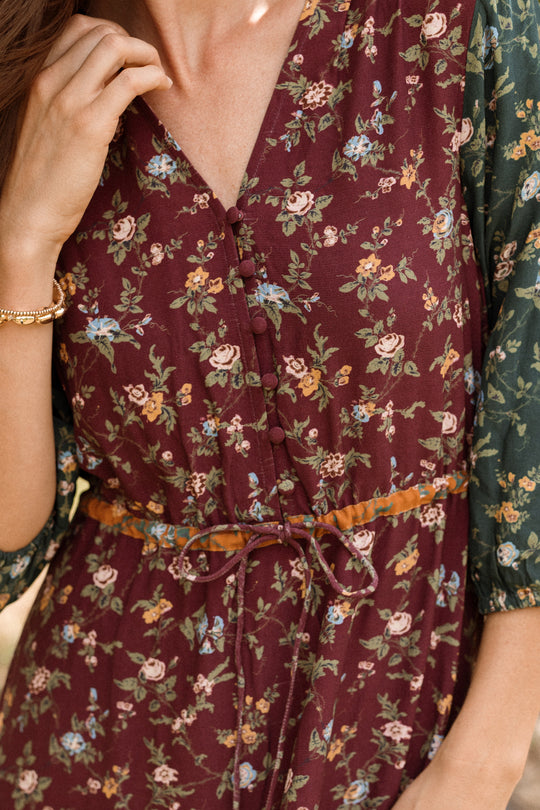 Close-up of a woman wearing the Harlowe Dress in Elysian Vine by Ivy City Co—a burgundy floral piece featuring a button front, drawstring waist, and green floral puff sleeves. A gold bracelet is visible on her wrist. This style offers a maternity-friendly design.