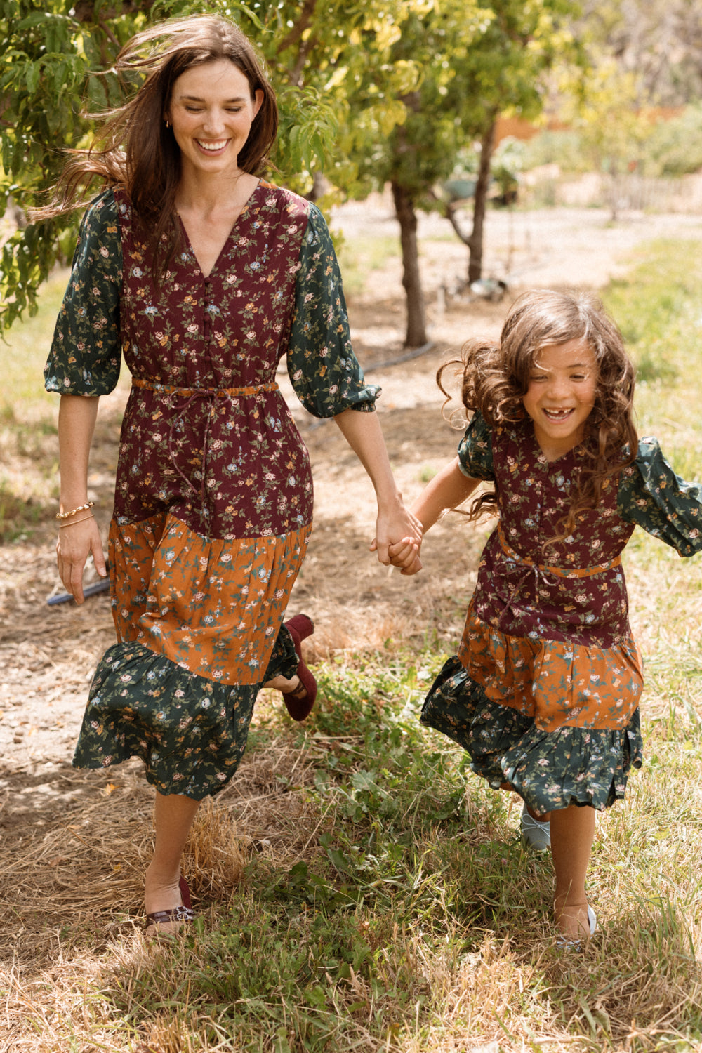 A smiling woman and young girl hold hands while running outdoors, both wearing matching Ivy City Co Harlowe Dresses in Elysian Vine. The belted, tiered floral dresses feature charming puff sleeves in a rich mix of burgundy, orange, and dark green hues and are maternity-friendly for effortless style at every stage.