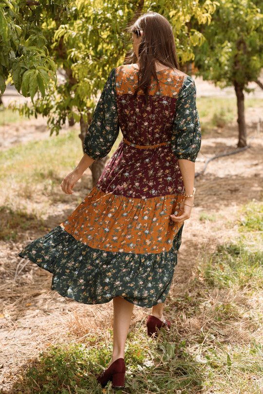 A woman, her long hair flowing freely and her back to the camera, strolls through a sunlit orchard in the Harlowe Dress in Elysian Vine from Ivy City Co. The puff sleeves and patchwork floral design add elegance to her maternity-friendly look, completed with maroon shoes and sunglasses.