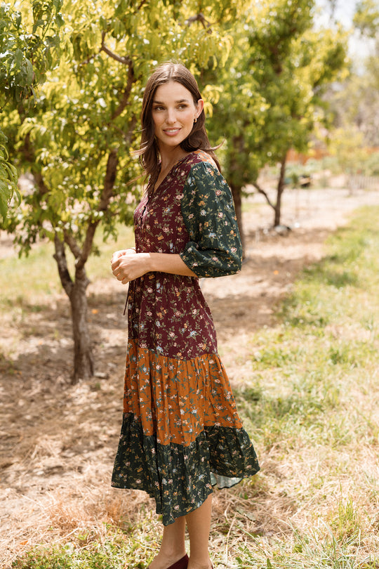 A woman wearing the Harlowe Dress in Elysian Vine by Ivy City Co, featuring puff sleeves and a floral patchwork design, stands outdoors among green trees, smiling softly in the sunshine.