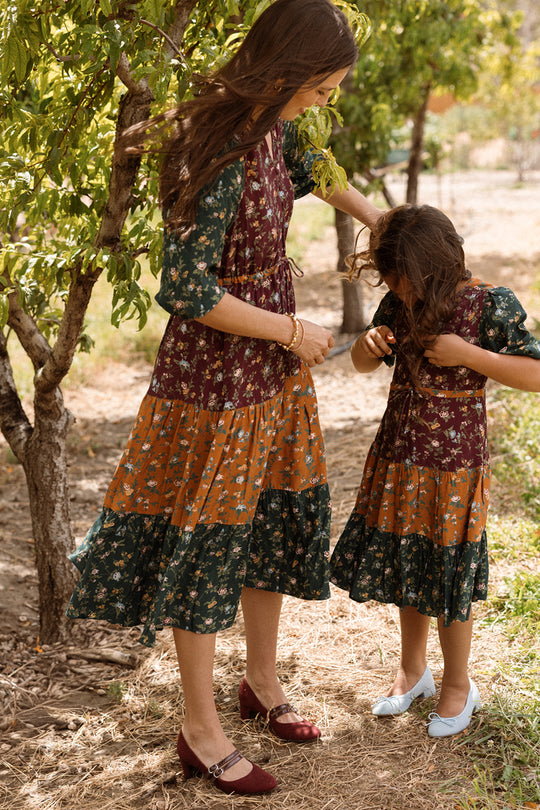 A woman and a girl, both wearing the Ivy City Co Harlowe Dress in Elysian Vine with charming puff sleeves, stand outdoors by the trees. The woman gently adjusts the girl’s hair as they look down, surrounded by greenery and sunlight. Their maternity-friendly dresses bring comfort to this serene moment.