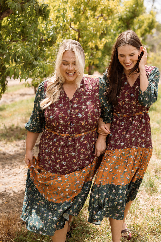 Two women wearing matching Ivy City Co Harlowe Dresses in Elysian Vine, featuring puff sleeves, walk arm-in-arm outdoors. They smile and laugh beneath green trees and warm sunlight. These stylish dresses are maternity friendly, making them perfect for every stage of life.