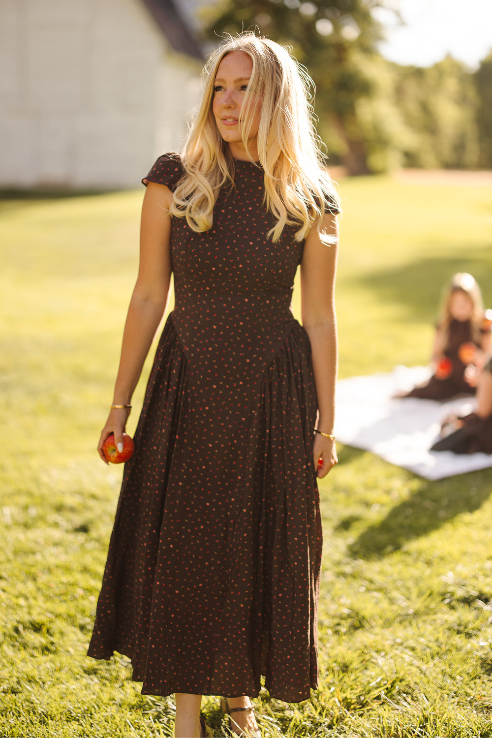 A woman in a brown, short-sleeved Harriet Dress in Chocolate Dainty Roses by Ivy City Co stands on grass holding an apple, with sunlight and blurred people on a picnic blanket in the background.