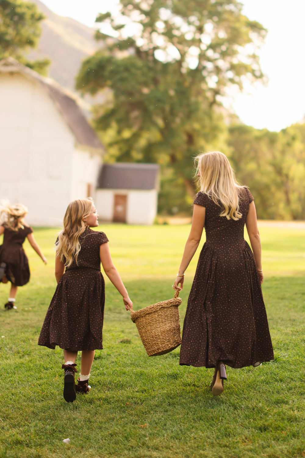 A woman and two girls, all dressed in Ivy City Co’s Harriet Dress in Chocolate Dainty Roses, walk together on the grass near a white barn. The woman carries a woven basket adorned with the Chocolate Dainty Roses print, while one girl walks at her side and the other runs ahead. They seem to be enjoying a sunny day outdoors.
