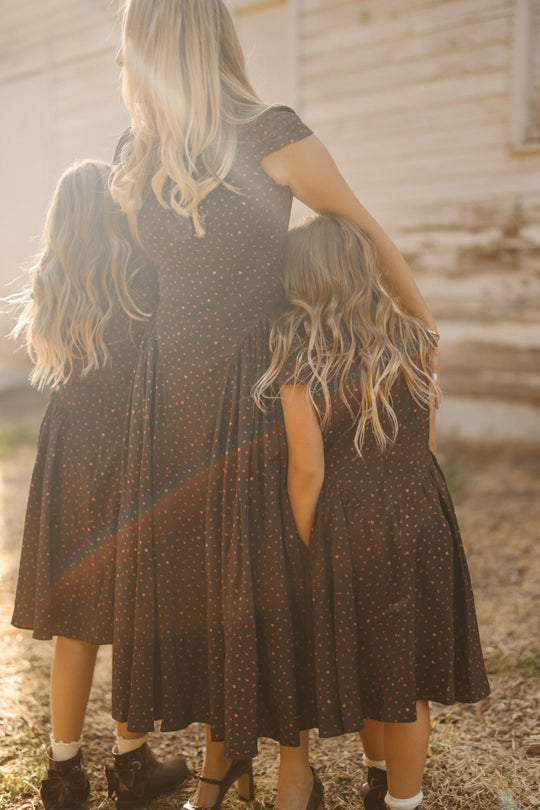 A woman and two young girls, all with long blonde hair and wearing matching Ivy City Co Harriet Dresses in Chocolate Dainty Roses, stand outdoors with their backs to the camera as sunlight shines on them.