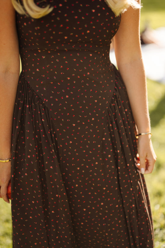 A woman wearing the Ivy City Co Harriet Dress in Chocolate Dainty Roses—a dark brown fall wedding dress adorned with a rich floral pattern—stands outdoors in sunlight, with only her torso and arms visible.