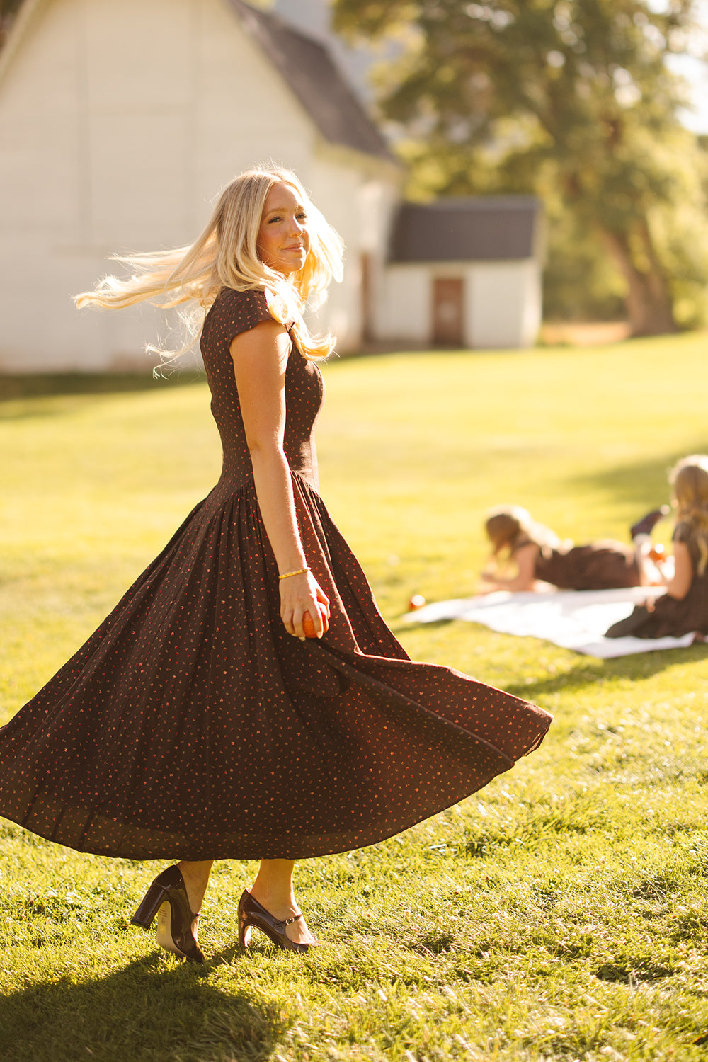 Dressed in Ivy City Co’s Harriet Dress in Chocolate Dainty Roses, a woman twirls on sunlit grass, her hair flowing. Nearby, two people relax on a picnic blanket decorated with chocolate dainty roses, set against the backdrop of a white building.