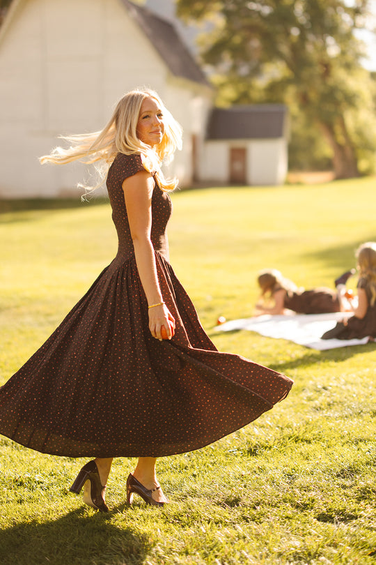 Dressed in Ivy City Co’s Harriet Dress in Chocolate Dainty Roses, a woman twirls on sunlit grass, her hair flowing. Nearby, two people relax on a picnic blanket decorated with chocolate dainty roses, set against the backdrop of a white building.