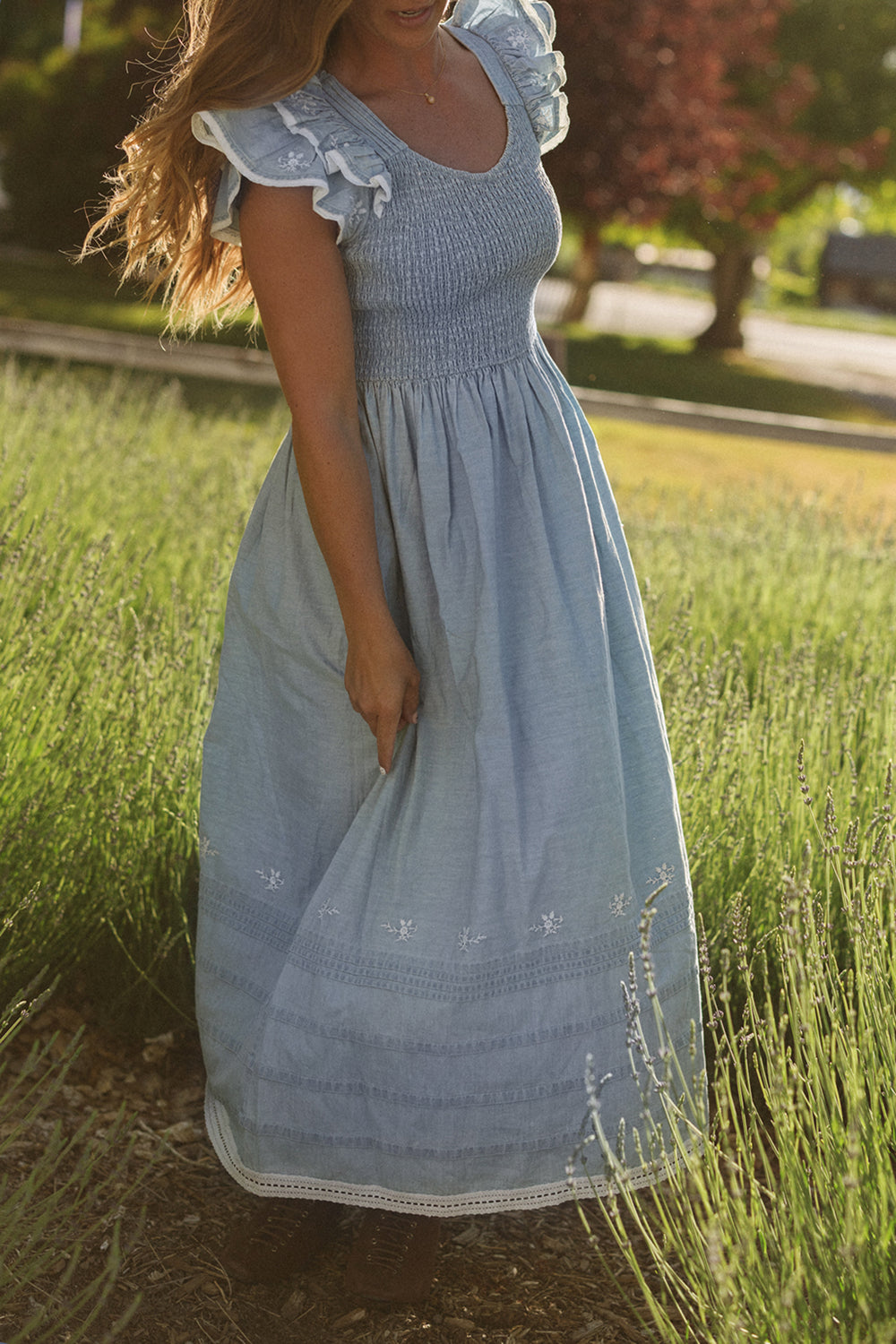 A woman in the Ivy City Co Hattie Ankle Length Dress in Embroidered Chambray, featuring ruffled sleeves and a delicate smocked bodice, stands outdoors on sunlit grass, holding the skirt of her maternity-friendly dress.