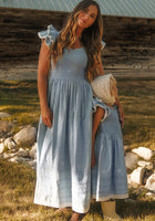 A woman and young child stand near a rustic barn, both wearing the Hattie Ankle Length Dress in Embroidered Chambray by Ivy City Co. These coordinated blue dresses feature ruffle sleeves, a smocked bodice, and white embroidered hems. Elegantly maternity friendly, this style is perfect for mothers and their little ones to match outdoors.