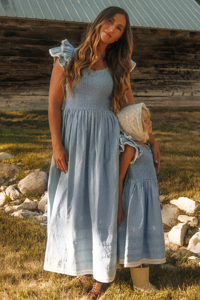 A woman and young child stand near a rustic barn, both wearing the Hattie Ankle Length Dress in Embroidered Chambray by Ivy City Co. These coordinated blue dresses feature ruffle sleeves, a smocked bodice, and white embroidered hems. Elegantly maternity friendly, this style is perfect for mothers and their little ones to match outdoors.