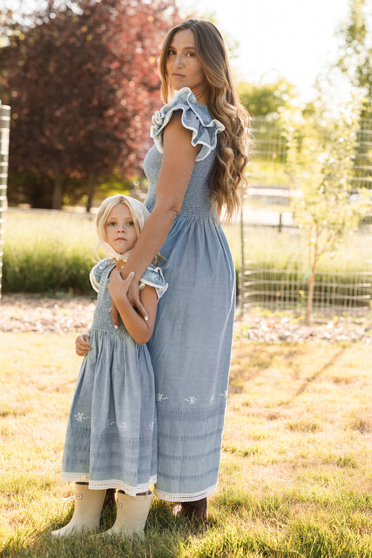 A woman and a young girl stand outdoors in golden sunlight, both wearing matching Ivy City Co Hattie Ankle Length Dresses in Embroidered Chambray with smocked bodices, ruffle sleeves, and white embroidered hems. The effortlessly elegant style is also maternity friendly.