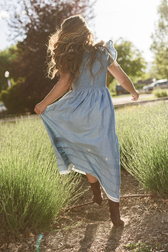A girl with long wavy hair wearing the Ivy City Co Hattie Ankle Length Dress in Embroidered Chambray, which is maternity friendly, runs down a path through tall grass on a sunny day. She holds up the skirt with its smocked bodice and wears brown boots.