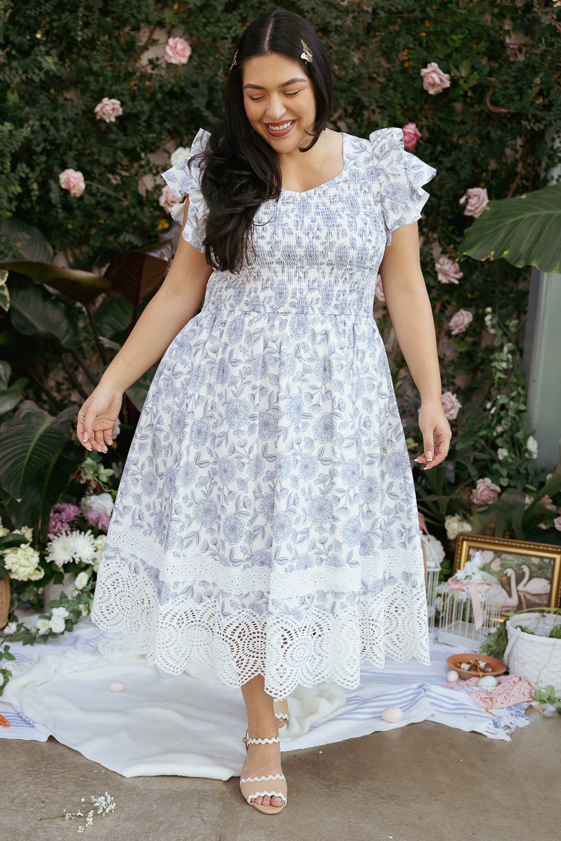 A woman with long dark hair smiles outdoors on a picnic blanket, wearing the Ivy City Co Hattie Dress in Blue Floral, surrounded by flowers and greenery.