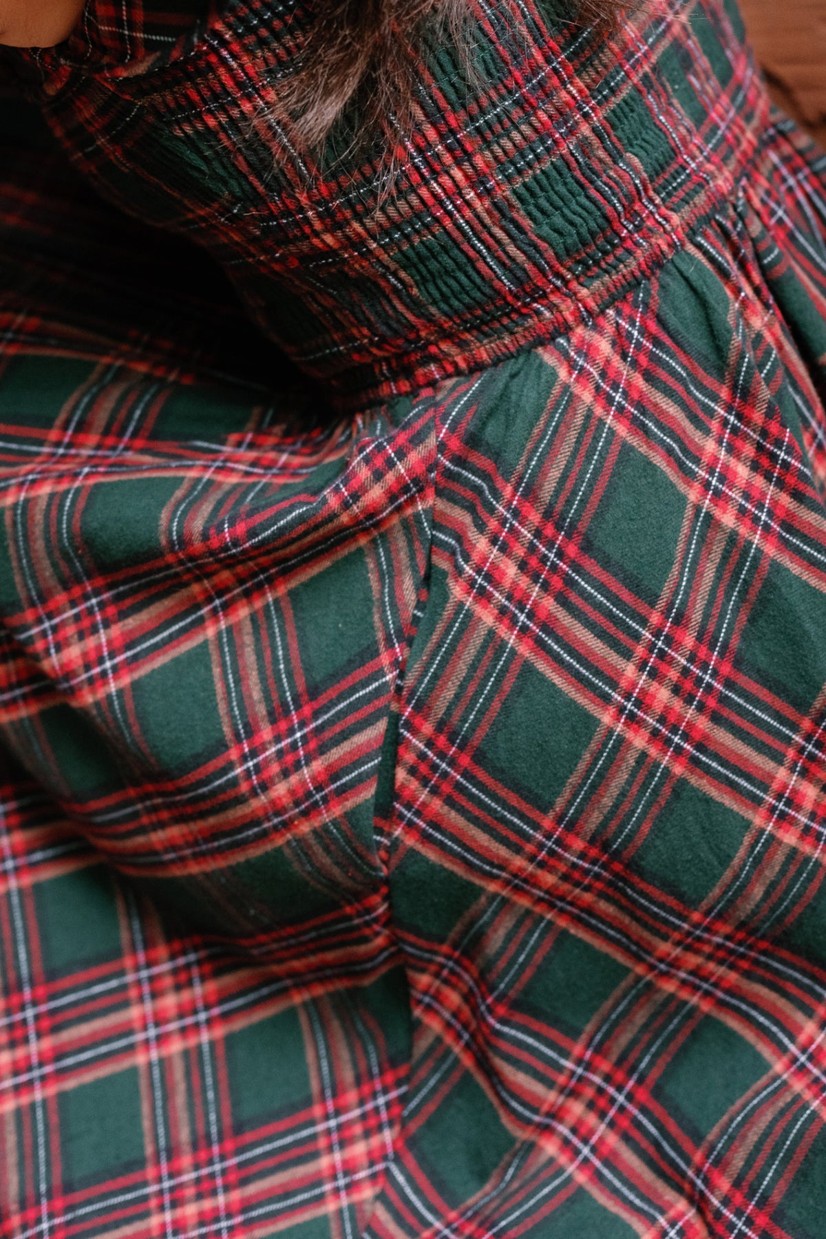 Close-up of the Hattie Dress in Evergreen Plaid by Ivy City Co, featuring a green fabric with red and white lines, highlighted by gathered stitching and pleats near the waist—giving this stylish dress a maternity-friendly fit ideal for any holiday occasion. A bit of dark hair is visible at the top edge.