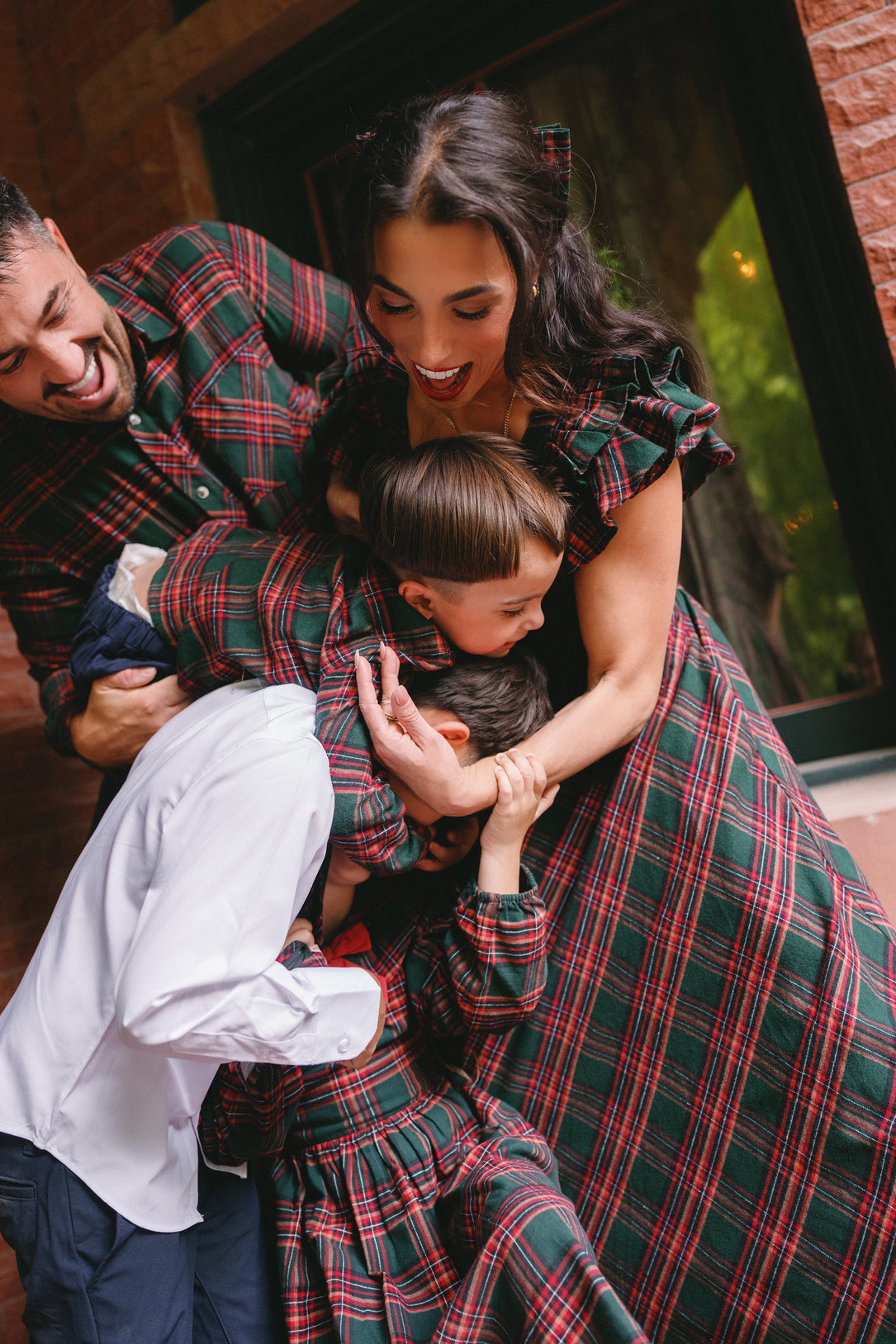 A joyful family in coordinated plaid outfits shares laughter and tight hugs in front of a brick building, with the mother wearing the Hattie Dress in Evergreen Plaid from Ivy City Co—a stylish and maternity-friendly holiday option. The playful moment is filled with warmth and energy.