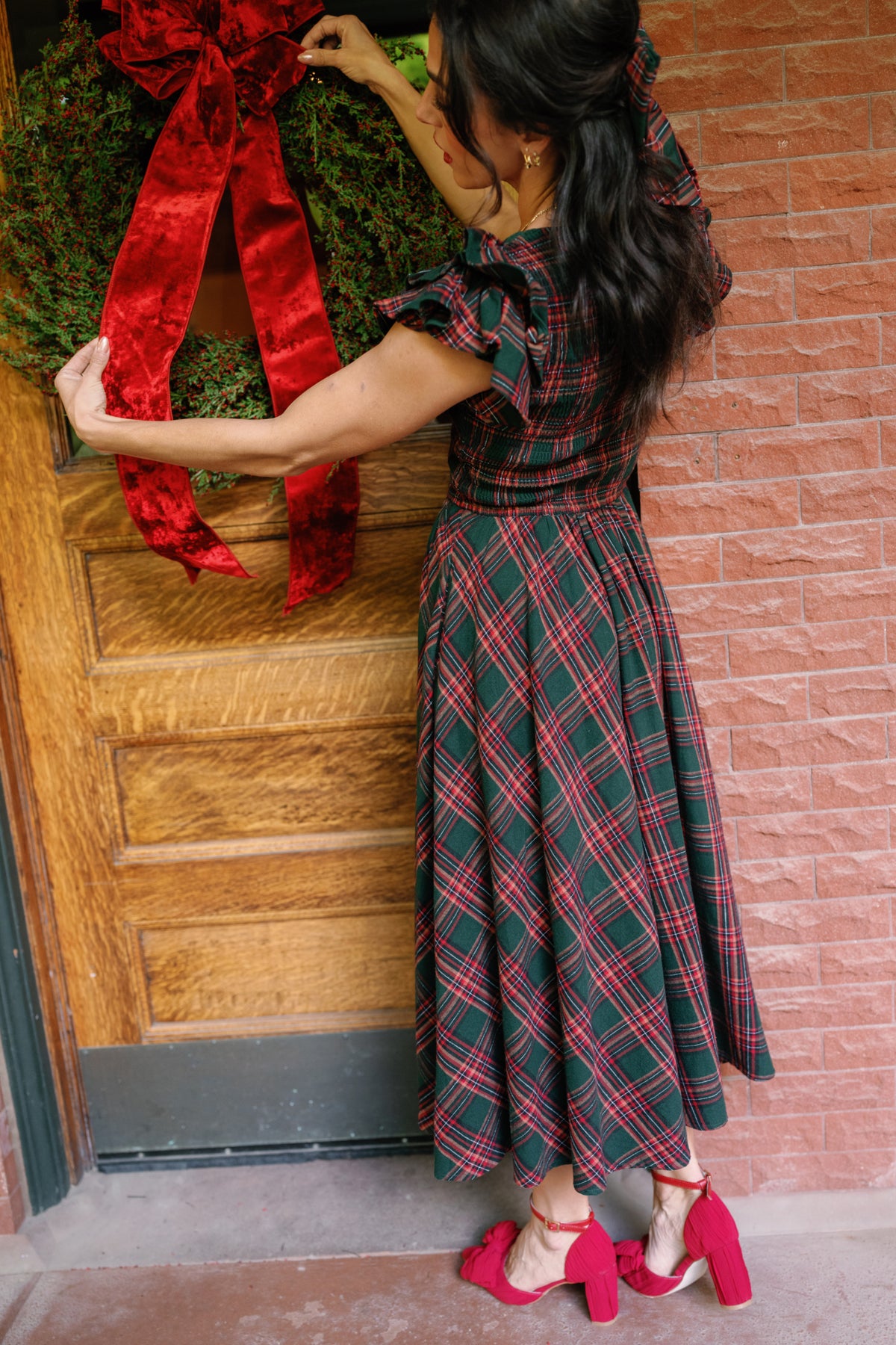 A woman with dark hair, wearing the Ivy City Co Hattie Dress in Evergreen Plaid paired with a matching hair bow and red block-heeled shoes, hangs a Christmas wreath adorned with a large red velvet bow on a wooden door beside a brick wall. This festive holiday dress is also maternity friendly, making it perfect for joyful gatherings.