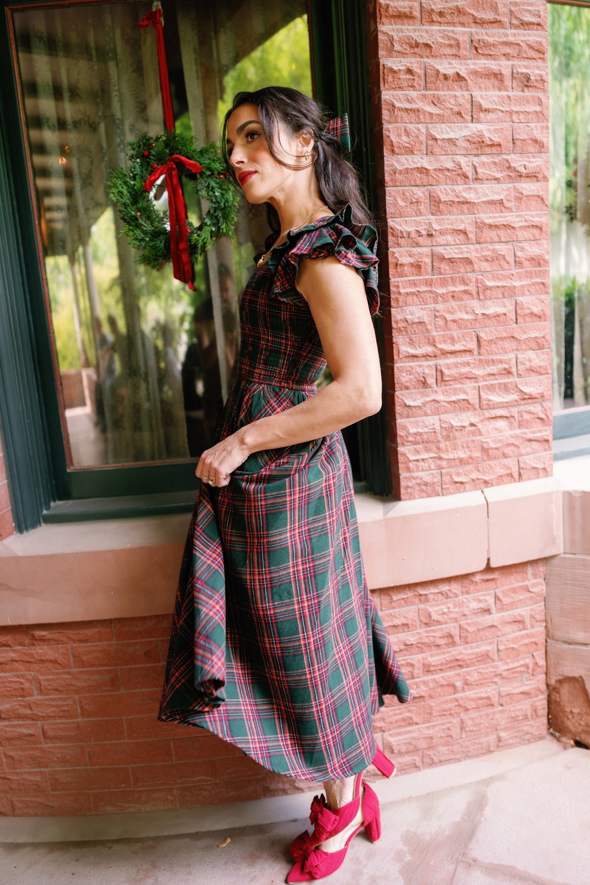 A woman wearing the Ivy City Co Hattie Dress in Evergreen Plaid and red shoes stands beside a brick wall and window decorated with a holiday wreath. She gazes to the side, holding out her maternity-friendly dress slightly, surrounded by festive décor.